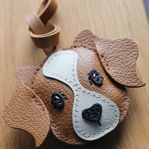 May include: A brown leather dog keychain with white and black details. The dog has stitched eyes, a nose, and ears.