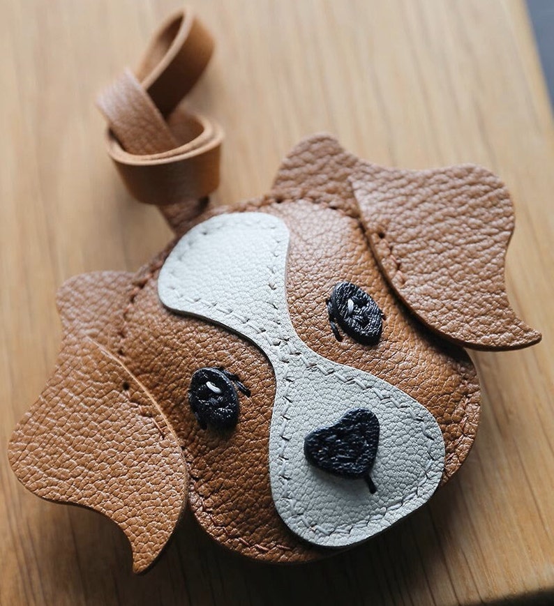 May include: A brown leather dog keychain with white and black details. The dog has stitched eyes, a nose, and ears.