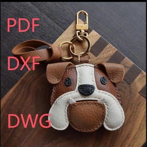Puede incluir: Un llavero de bulldog de cuero marrón, blanco y negro con un cierre y un llavero dorados. El llavero tiene una correa de cuero marrón y las letras PDF, DXF y DWG en rojo.