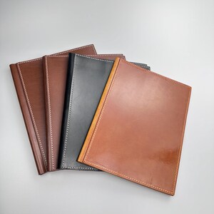 May include: A collection of leather journals in various shades of brown and black. Each journal features a smooth, glossy finish and is accented with contrasting white or cream stitching around the edges. The journals are arranged in a fan-like display.