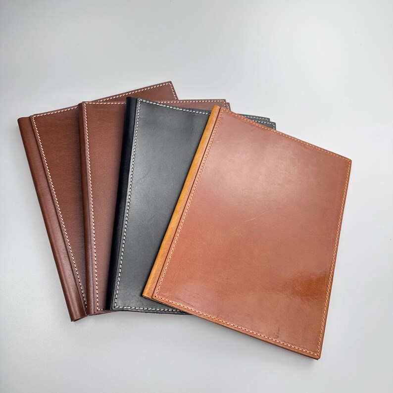 May include: A collection of leather journals in various shades of brown and black. Each journal features a smooth, glossy finish and is accented with contrasting white or cream stitching around the edges. The journals are arranged in a fan-like display.