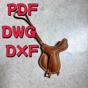 Peut inclure: Une petite selle en cuir marron avec des coutures blanches et une boucle en métal. La selle a une boucle en haut et est présentée sur un fond gris. L'image comprend également le texte "PDF", "DWG" et "DXF" en rouge.