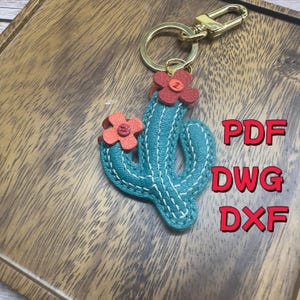 Puede incluir: Un llavero en forma de cactus turquesa con detalles de flores rojas y costuras blancas. El llavero tiene un cierre y un anillo dorados. El texto a la derecha dice "PDF", "DWG" y "DXF".