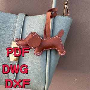 Puede incluir: Un charm de bolso con forma de perro de cuero marrón con costuras blancas, colgando de un bolso verde claro. El charm tiene un lazo para sujetarlo. Las palabras "PDF", "DWG" y "DXF" están impresas en rojo.