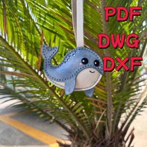Może przedstawiać: Ręcznie robiona niebieska ozdoba w kształcie wieloryba z kremowym brzuchem i szytymi detalami. Wieloryb ma czarne oczy i uśmiechnięte usta. Tekst "PDF, DWG, DXF" jest wyświetlany na czerwono.