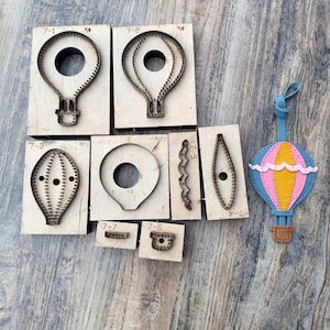 May include: A collection of wooden die-cut tools for crafting, featuring various shapes of hot air balloons. The set includes several balloon outlines, a decorative edge, and a finished leather hot air balloon with pink, yellow, and blue colors.