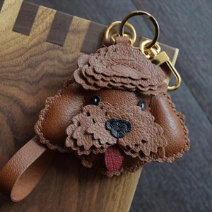 Puede incluir: Llavero de cuero marrón con forma de cara de perro. El perro tiene una nariz negra y una lengua roja. El llavero tiene herrajes dorados y una correa de cuero marrón.