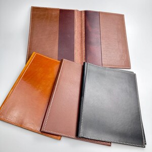 May include: A collection of leather journals and covers in various shades of brown and black. The journals are open and closed, showcasing the interior pages and the stitched edges. The leather has a glossy finish.