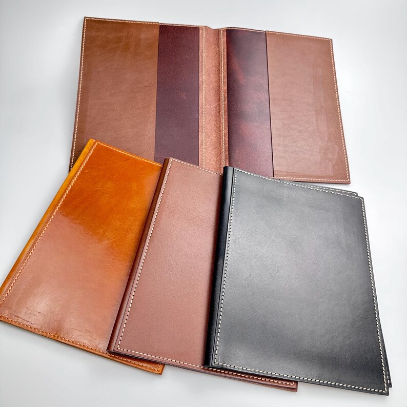 May include: A collection of leather journals and covers in various shades of brown and black. The journals are open and closed, showcasing the interior pages and the stitched edges. The leather has a glossy finish.