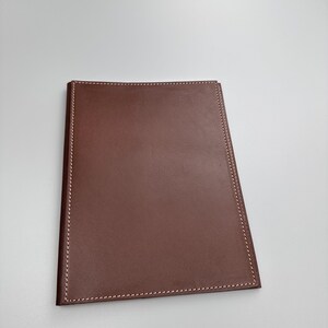 May include: A brown leather portfolio with white stitching around the edges. The portfolio is rectangular with a slightly trapezoidal shape. The leather has a smooth texture and a rich, warm color.