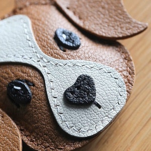 May include: Close-up of a brown and white leather dog keychain with black stitched details. The dog has a heart-shaped nose and two black eyes.