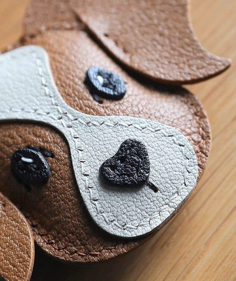 May include: Close-up of a brown and white leather dog keychain with black stitched details. The dog has a heart-shaped nose and two black eyes.