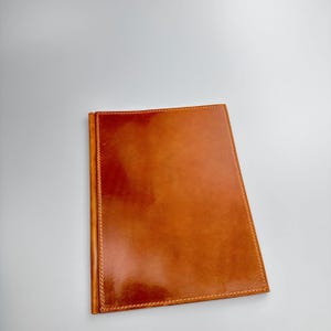 May include: A rectangular, brown leather journal with a stitched border. The cover has a glossy finish and a warm, reddish-brown hue. The journal is closed and appears to be new.