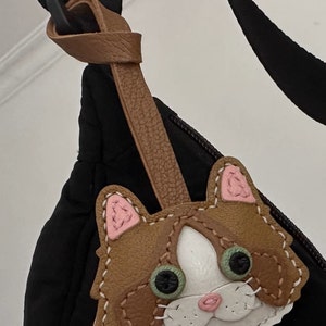May include: A brown leather cat keychain with pink ears, white fur, and green eyes. The keychain is attached to a brown leather strap.