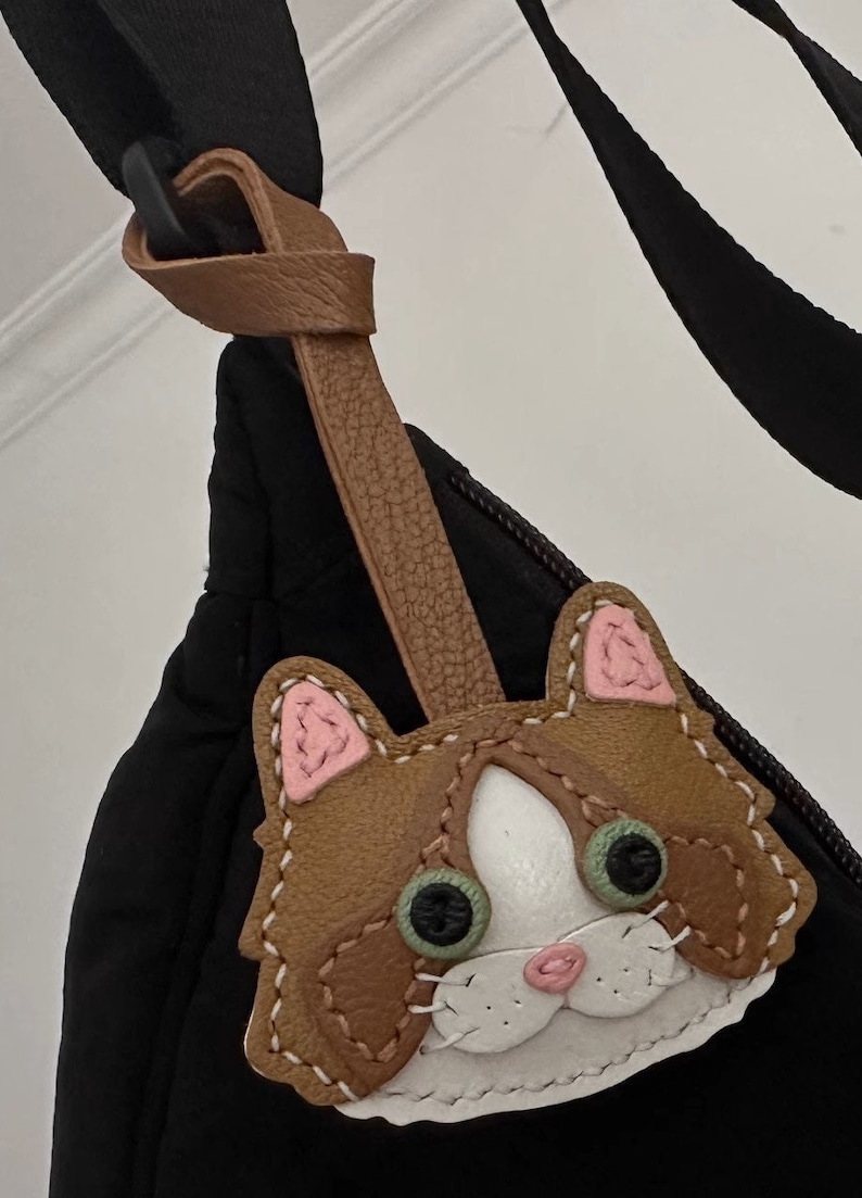 May include: A brown leather cat keychain with pink ears, white fur, and green eyes. The keychain is attached to a brown leather strap.