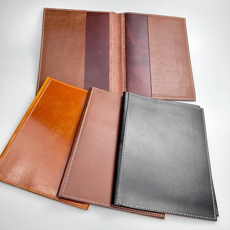 May include: A collection of leather journals and covers in various shades of brown and black. The journals are rectangular with white stitching along the edges. One journal is open, revealing a multi-toned interior. The leather has a polished finish.
