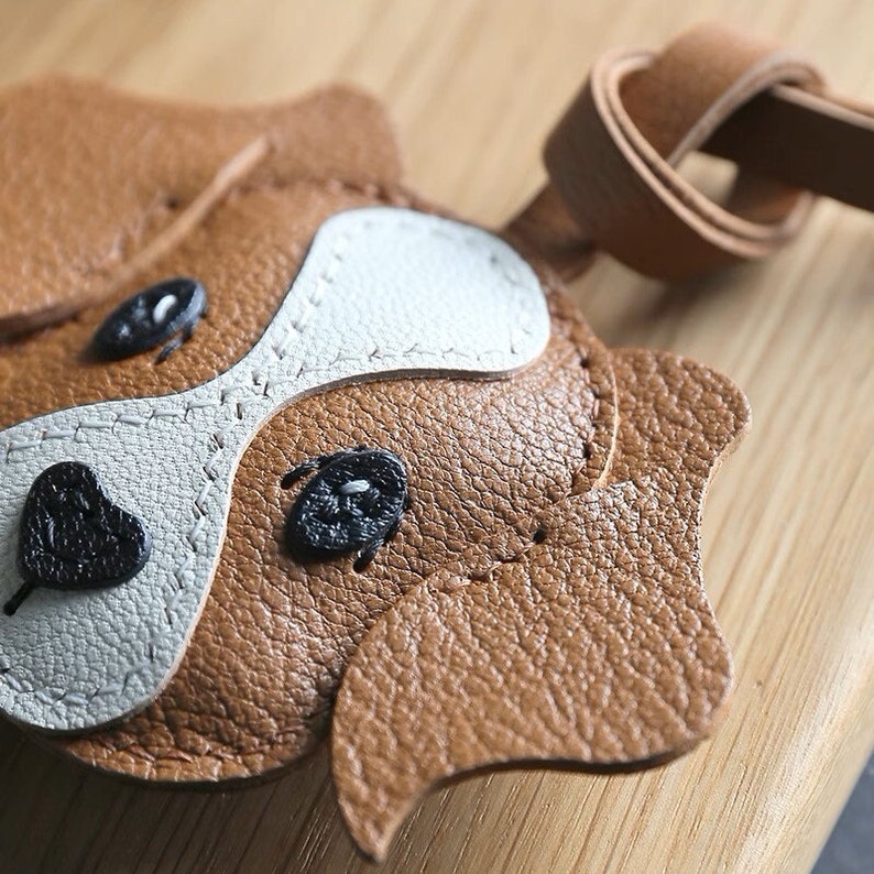 May include: A brown leather dog keychain with white and black accents. The dog's face is stitched with detail and has black button eyes.