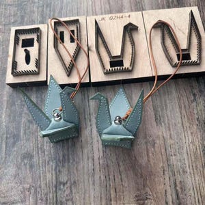 May include: Two light blue leather crane ornaments with brown leather cords, next to wooden die-cut shapes. The crane ornaments have silver-colored metal accents and white stitching. The wooden shapes are for leather crafting.