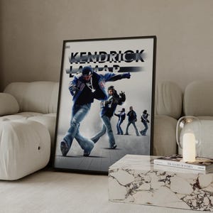Op de afbeelding: Een ingelijste poster met een afbeelding van Kendrick Lamar in een blauwe jas en jeans, met de naam van de artiest in het wit. Het kunstwerk toont Lamar in beweging, met andere figuren op de achtergrond, wat een dynamische compositie creëert.