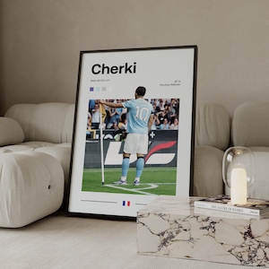 May include: Framed poster featuring a soccer player in a light blue jersey with the number 10. The poster has the name "Cherki" in bold letters, with "Manchester City" below. The player is on a green field, holding a flag. The poster is in a black frame.