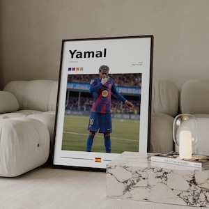 May include: Framed poster featuring the name "Yamal" in bold black letters. The poster displays a soccer player in a blue and red uniform. The Spanish flag is at the bottom. The poster is displayed on a white marble table.