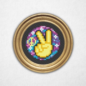 May include: A framed cross-stitch artwork showing a yellow peace sign hand gesture. The hand is surrounded by a colourful design of stars and a peace symbol, set against a black background. The frame is gold-coloured.