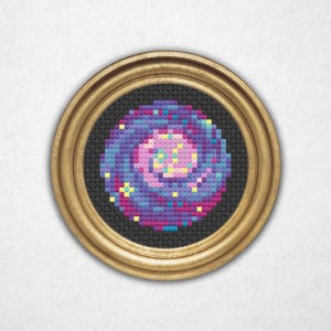 May include: A framed cross-stitch embroidery depicting a swirling galaxy with a pink center and yellow stars. The galaxy is surrounded by a dark blue background.