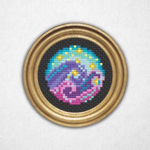 Puede incluir: Una bordadura de punto de cruz enmarcada que representa una galaxia giratoria en colores rosa, morado y azul. La galaxia está rodeada por un anillo de estrellas amarillas sobre un fondo negro.