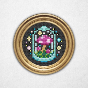 May include: A cross-stitch artwork of a mushroom terrarium in a round, gold-coloured frame. The mushroom has a pink cap with white spots, and is surrounded by green foliage and colourful accents. The background is black.
