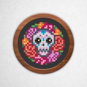 Può includere: Un disegno a punto croce rotondo con un teschio di zucchero circondato da una ghirlanda floreale. Il teschio è bianco con accenti colorati e i fiori sono nelle tonalità del rosa, dell'arancione e del verde. Il disegno è su uno sfondo grigio scuro e incorniciato da un bordo in legno.