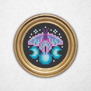 Puede incluir: Una obra de arte de punto de cruz enmarcada con un diseño de mariposa estilizada. La mariposa está representada en tonos de rosa, morado y turquesa sobre un fondo negro. La obra está encerrada en un marco redondo de color dorado.