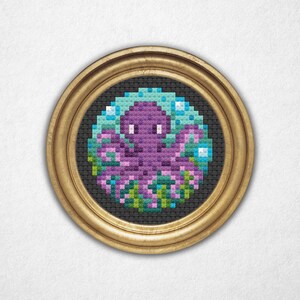 May include: A framed cross-stitch artwork featuring a purple octopus surrounded by blue bubbles and pink and green plants. The artwork is set against a black background and framed in a gold-colored, round frame. The piece is displayed on a white background.