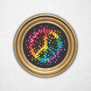 May include: A framed, round cross-stitch piece featuring a colorful peace sign. The sign is composed of pixelated squares in a rainbow of colors against a black background. The frame is a warm gold color, and the piece is centered.