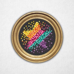 Puede incluir: Una obra de arte de punto de cruz enmarcada, de forma redonda. El diseño presenta un patrón abstracto pixelado y colorido en tonos de amarillo, naranja, rosa, turquesa y morado sobre un fondo negro. El marco es dorado.