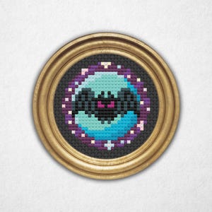 May include: A framed cross-stitch artwork with a pixelated design of a teal and blue orb and a black central shape, surrounded by purple and white accents. The artwork is set within a round, gold-coloured frame.
