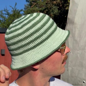Könnte beinhalten: Gehäkelter Fischerhut mit grünen und weißen Streifen. Der Hut hat eine runde Krone und eine hochgebogene Krempe. Die Person im Bild trägt ein weißes T-Shirt. Ein modisches Accessoire.