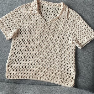 May include: A cream-colored, short-sleeved crochet shirt with a collared neckline. The shirt features a V-neck and a loose, open weave design. The garment is made of a soft, natural-looking yarn.