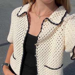 Könnte beinhalten: Cremefarbene Häkeljacke mit schwarzem Besatz. Die kurzärmelige Jacke hat einen Kragen, zwei Vordertaschen und goldfarbene Knöpfe. Das offene Webdesign ist im gesamten Kleidungsstück sichtbar. Ein stilvoller Zusatz für jede Garderobe.