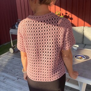 May include: A light pink, short-sleeved crochet top with a collared neckline. The open weave design of the top allows for breathability. The top is worn with a black skirt.