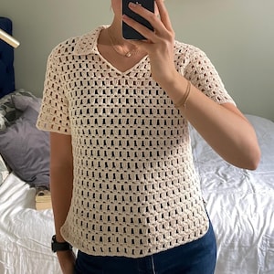 May include: Cream-colored crochet polo shirt with short sleeves. The shirt features a V-neck collar and a loose, open weave design. The shirt is made of a soft, breathable material, perfect for warm weather. A stylish and comfortable addition to any wardrobe.