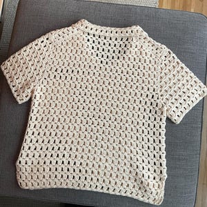 May include: A cream-colored, short-sleeved crochet top. The open weave design creates a grid-like pattern throughout the shirt. The top has a collared neckline and a relaxed fit, perfect for layering or wearing on its own.