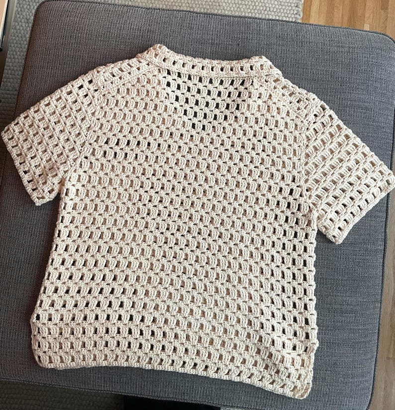 May include: A cream-colored, short-sleeved crochet top. The open weave design creates a grid-like pattern throughout the shirt. The top has a collared neckline and a relaxed fit, perfect for layering or wearing on its own.