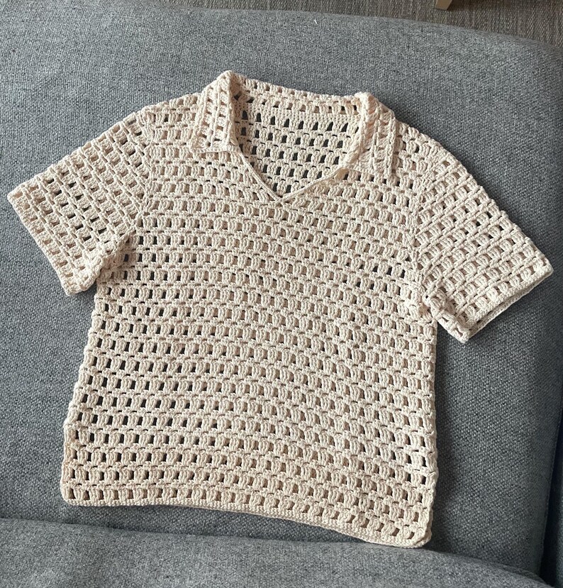 May include: A cream-colored, short-sleeved crochet shirt with a collared neckline. The shirt features a V-neck and a loose, open weave design. The garment is made of a soft, natural-looking yarn.