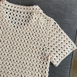 May include: Cream-colored crochet top with short sleeves. The open weave design creates a textured look. The collar and sleeves have a more solid, defined edge. The top is made of a soft, natural-looking yarn.