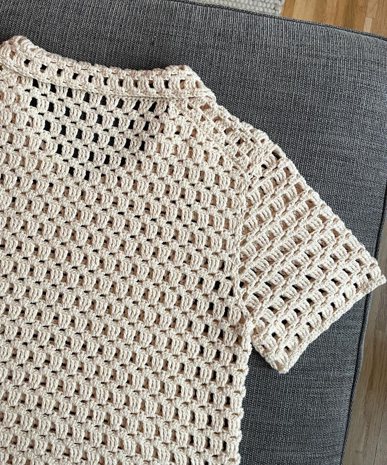 May include: Cream-colored crochet top with short sleeves. The open weave design creates a textured look. The collar and sleeves have a more solid, defined edge. The top is made of a soft, natural-looking yarn.