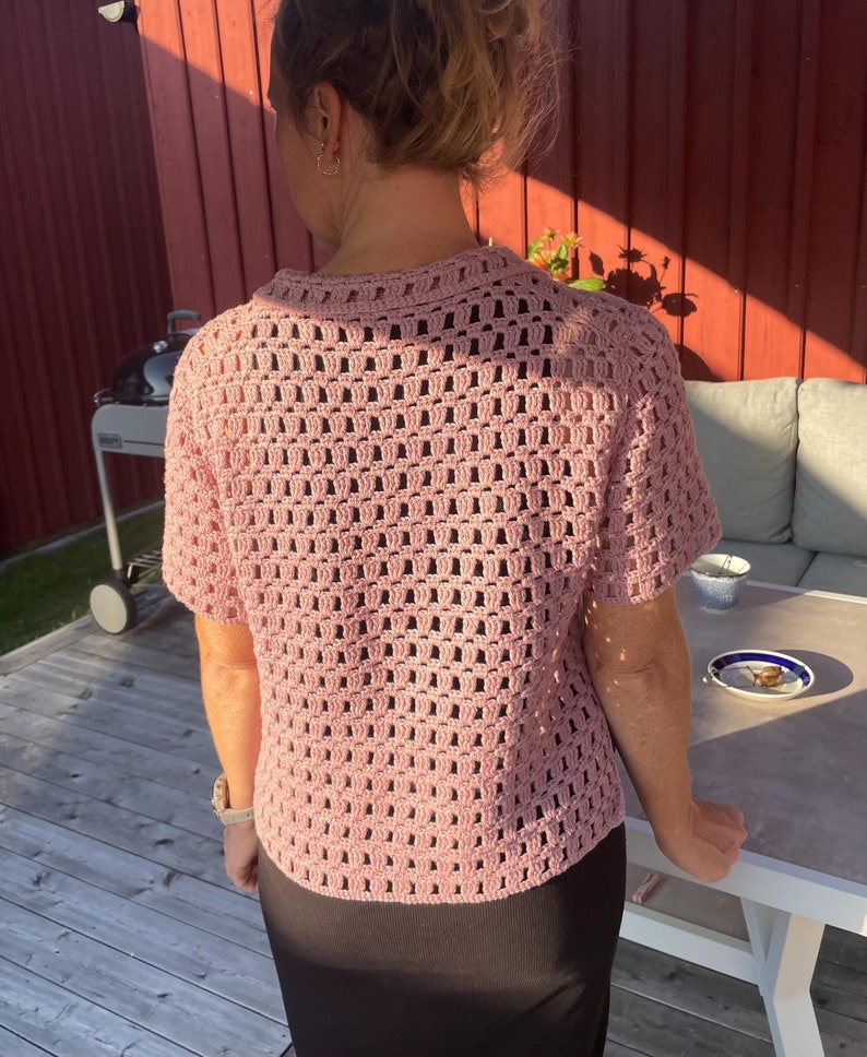 May include: A light pink, short-sleeved crochet top with a collared neckline. The open weave design of the top allows for breathability. The top is worn with a black skirt.