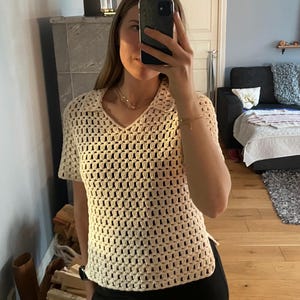 May include: Cream-colored, open-weave crochet top with short sleeves and a V-neck collar. The top is paired with black pants. The image is taken indoors, with a mirror reflection showing the person wearing the top.