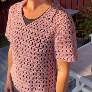 May include: A short-sleeved, light pink crochet top with a V-neck collar. The open weave design of the shirt is visible. The top is worn over a dark-colored undershirt. The shirt is a handmade garment.