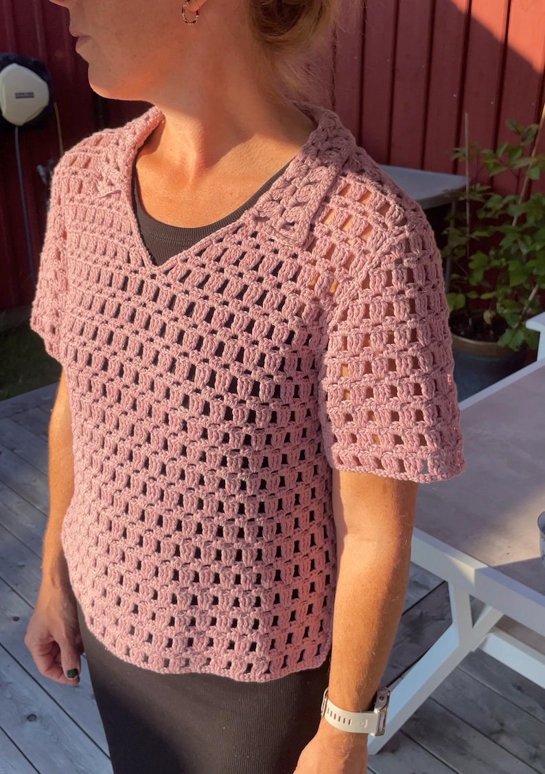 May include: A short-sleeved, light pink crochet top with a V-neck collar. The open weave design of the shirt is visible. The top is worn over a dark-colored undershirt. The shirt is a handmade garment.