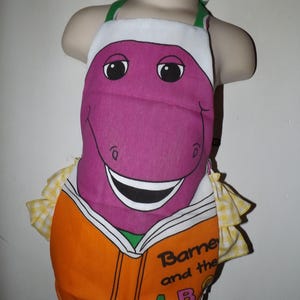 May include: A white apron featuring a large purple dinosaur character graphic holding an orange book with the text Barney and the ABCs. The apron has green neck straps and yellow and white checkered ruffled trim along the sides of the garment.